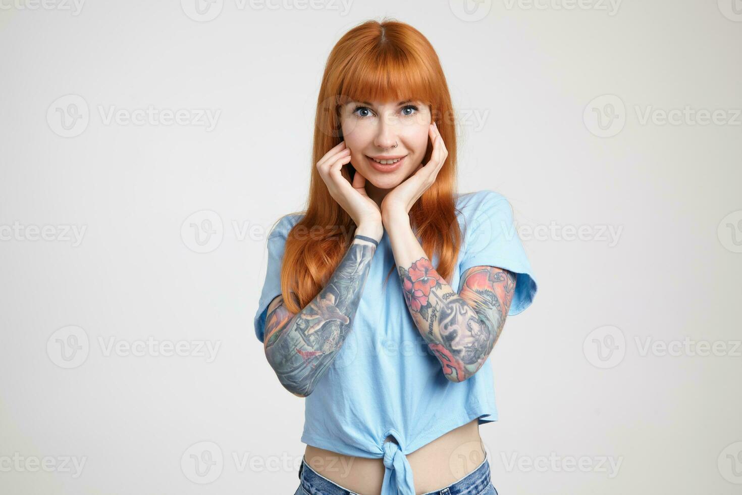 Indoor shot of young beautiful long haired redhead female holding her face with raised hands while smiling gladly at camera, isolated over white background photo