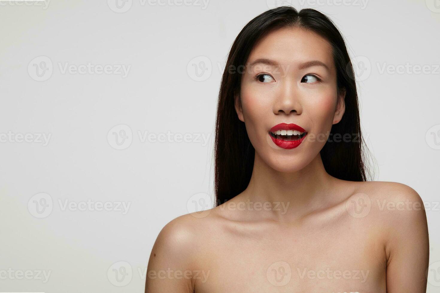 Studio photo of young beautiful dark haired woman with red lips looking excitedly aside and keeping her mouth opened, standing against white background
