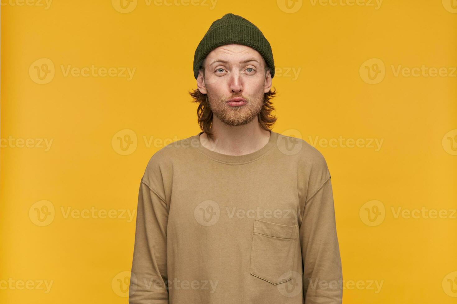 Cool looking male, handsome bearded guy with blond hairstyle. Wearing green beanie and beige sweater. Purses his lips in a kiss. Watching at the camera isolated over yellow background photo