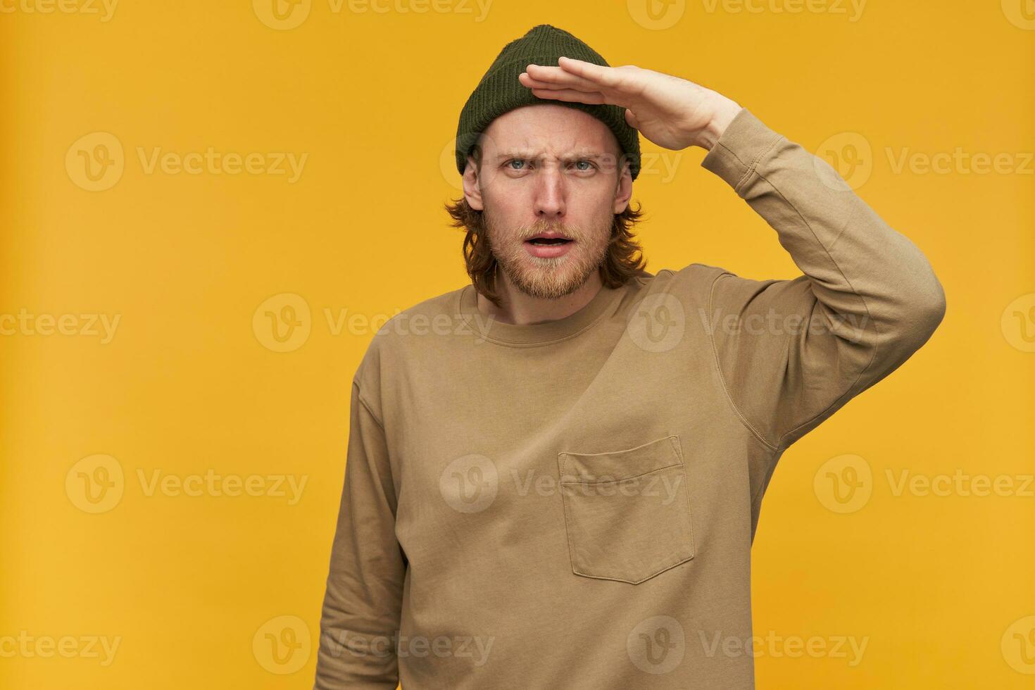 Portrait of serious male with blond hair and beard. Wearing green beanie and beige sweater. Holds palm next to forehead and gaze into distance. Watching at the camera isolated over yellow background photo
