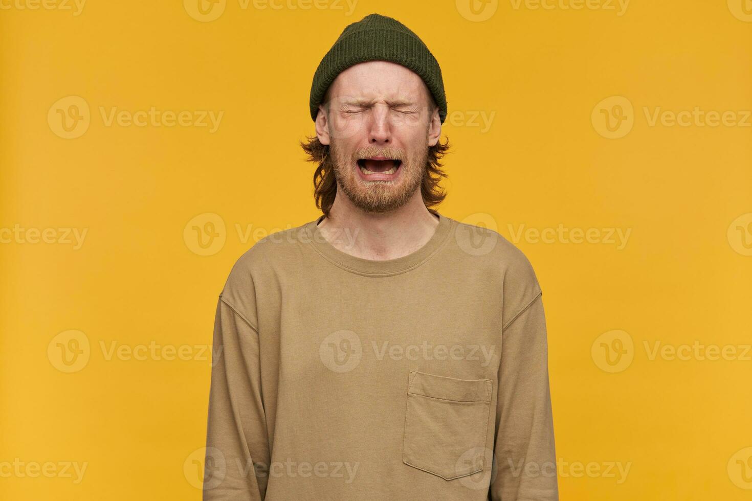 Despaired man, tearful guy with blond hair, beard and mustache. Wearing green beanie and beige sweater. Crying with closed eyes and feels hopeless. Stand isolated over yellow background photo