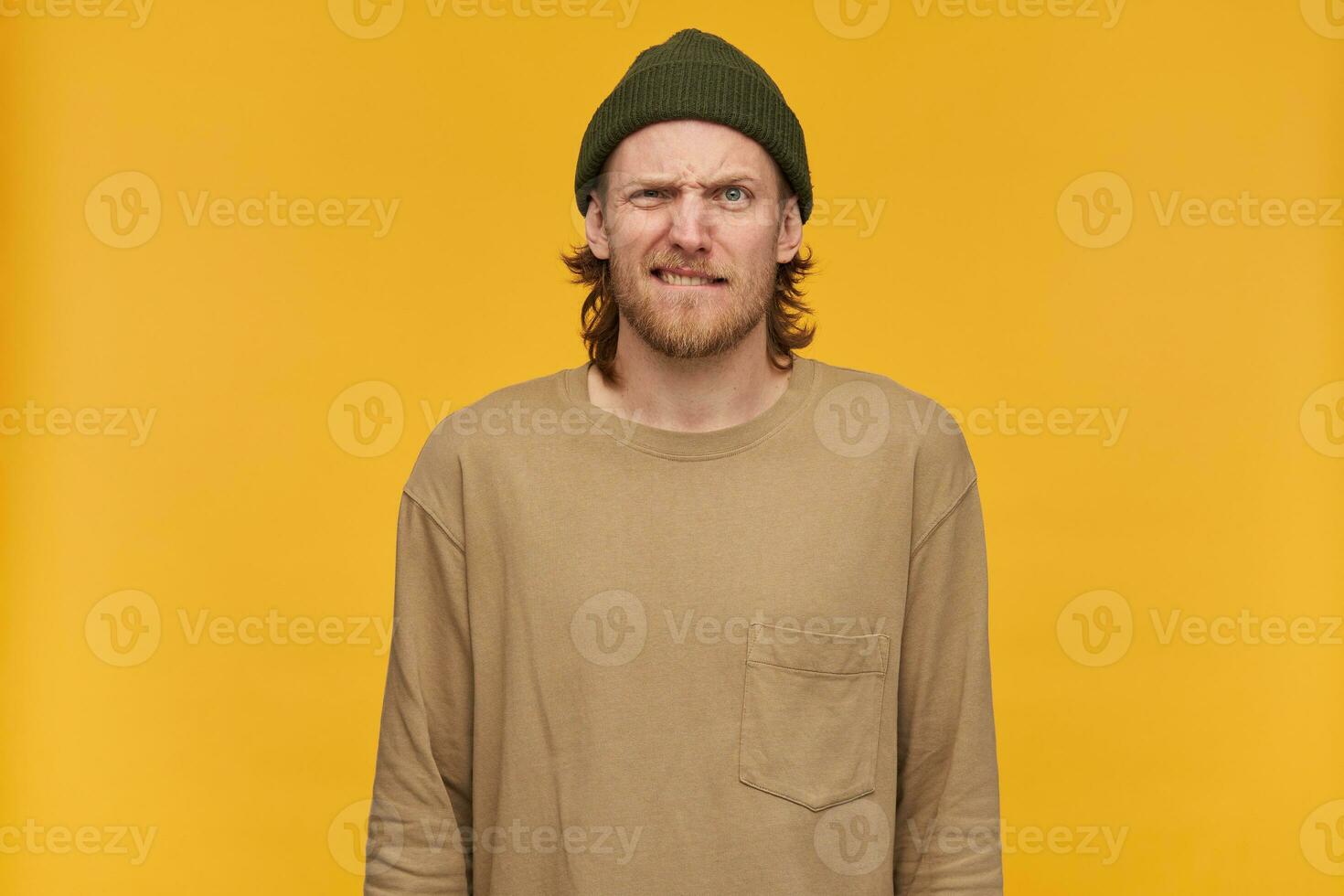 Angry looking male, frowning bearded guy with blond hairstyle. Wearing green beanie and beige sweater. Bites his lip. Watching at the camera isolated over yellow background photo