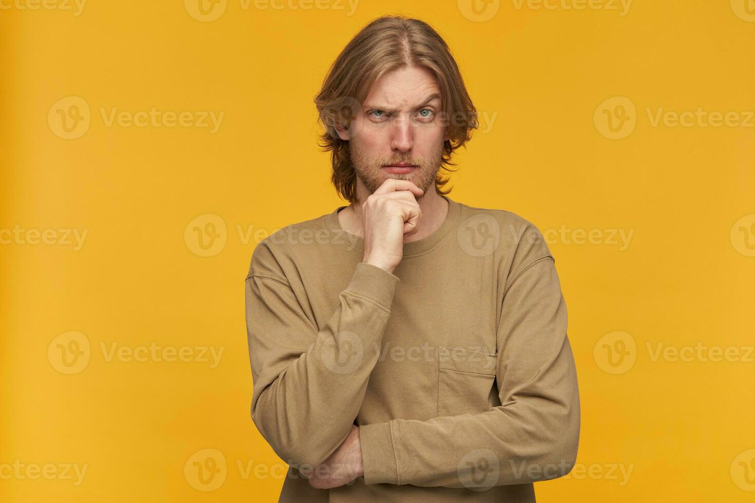 Young man, thoughtful guy with blond hair, beard and mustache. Wearing beige sweater. Touching his chin and lifts an eyebrow. Watching at the camera isolated over yellow background photo