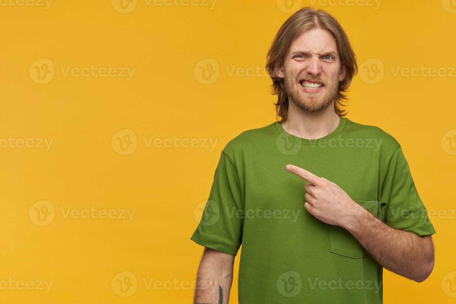 Irritated, unsure guy with blond hair, beard. Wearing green t-shirt. Has tattoo. Wry his face. Watching at the camera and pointing finger to the left at copy space, isolated over yellow background photo