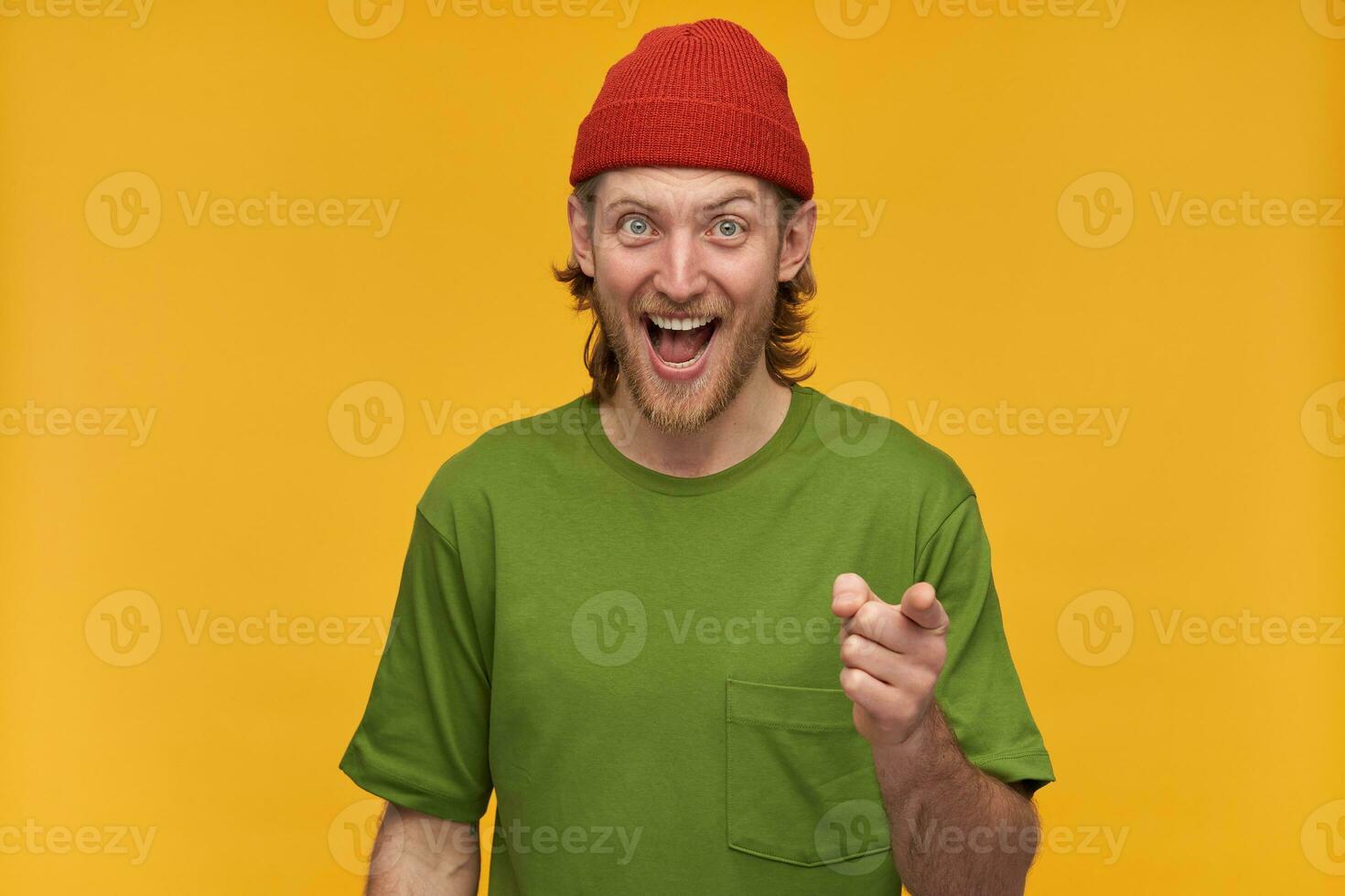 Portrait of cheerful, positive man with blond hairstyle and beard. Wearing green t-shirt and red beanie. Laughing and pointing finger at you. Watching at the camera isolated over yellow background photo