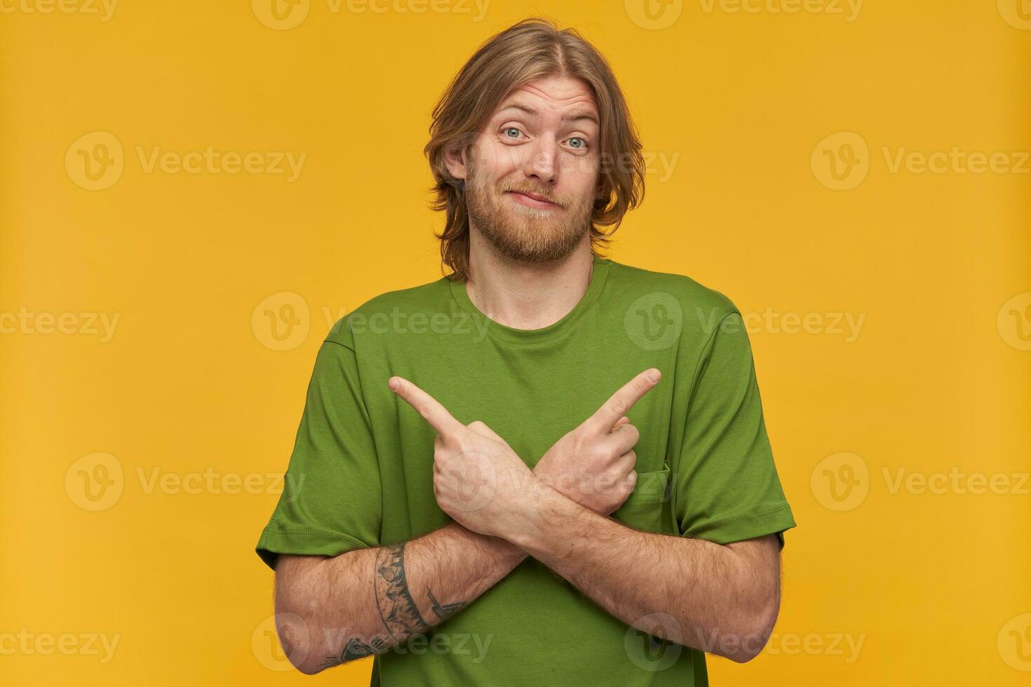 Confused, unsure bearded guy with blond hairstyle. Wearing green t-shirt. Has tattoo. Wry his face. Watching at the camera and pointing both sides at copy space, isolated over yellow background photo