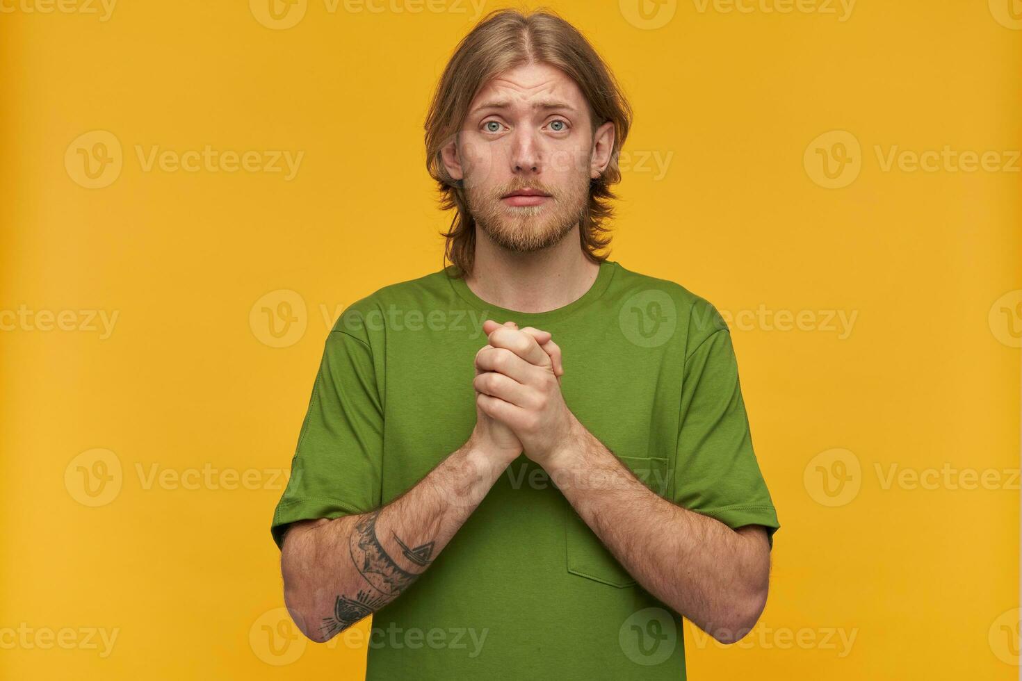 Orando masculino, mendicidad barbado chico con rubio peinado. vistiendo verde camiseta. tiene tatuajes sostiene palmas juntos y alegar. acecho a el cámara aislado terminado amarillo antecedentes foto