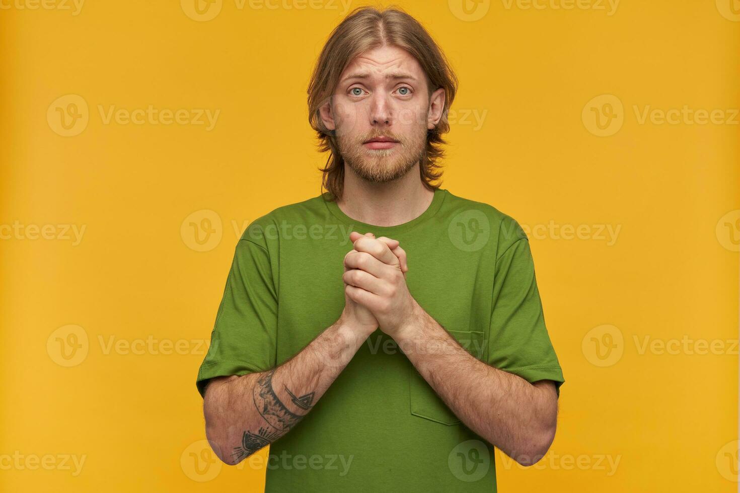 Praying male, begging bearded guy with blond hairstyle. Wearing green t-shirt. Has tattoos. Holds palms together and plead. Watching at the camera isolated over yellow background photo