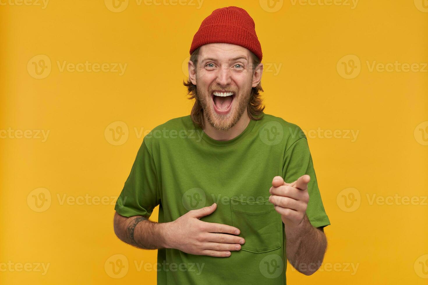 Portrait of positive, adult male with blond hairstyle and beard. Wearing green t-shirt and red beanie. Has tattoo. Laughing hard from you. Watching at the camera isolated over yellow background photo