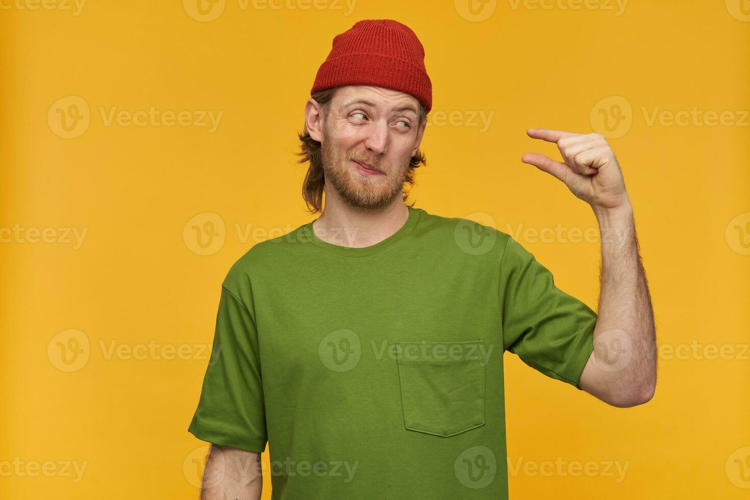 Unsure looking male, handsome bearded guy with blond hair. Wearing green t-shirt and red beanie. Showing little size and looking doubtedly at it. Stand isolated over yellow background photo