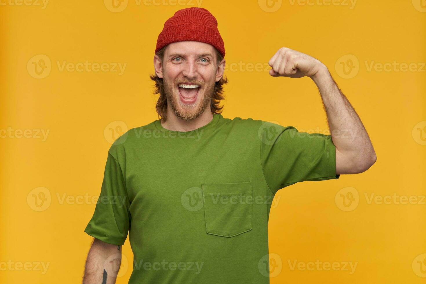 joven hombre, alegre chico con rubio cabello, barba y Bigote. vistiendo verde camiseta y rojo gorro. tiene tatuaje. muestra su bíceps, fortaleza. acecho a el cámara aislado terminado amarillo antecedentes foto