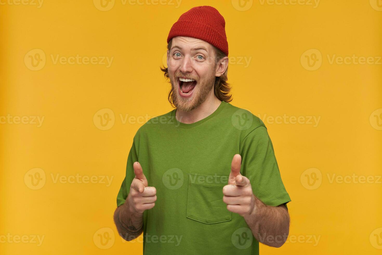 Portrait of positive, cheerful male with blond hairstyle and beard. Wearing green t-shirt and red beanie. Has tattoo. Pointing fingers at you. Watching at the camera isolated over yellow background photo