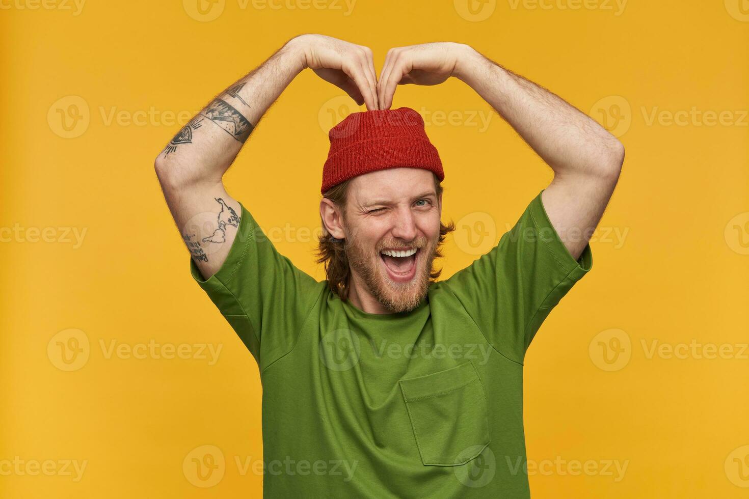 Portrait of handsome, adult male with blond hairstyle and beard. Wearing green t-shirt and red beanie. Has tattoos. Making heart sign with hands. Winks at the camera isolated over yellow background photo