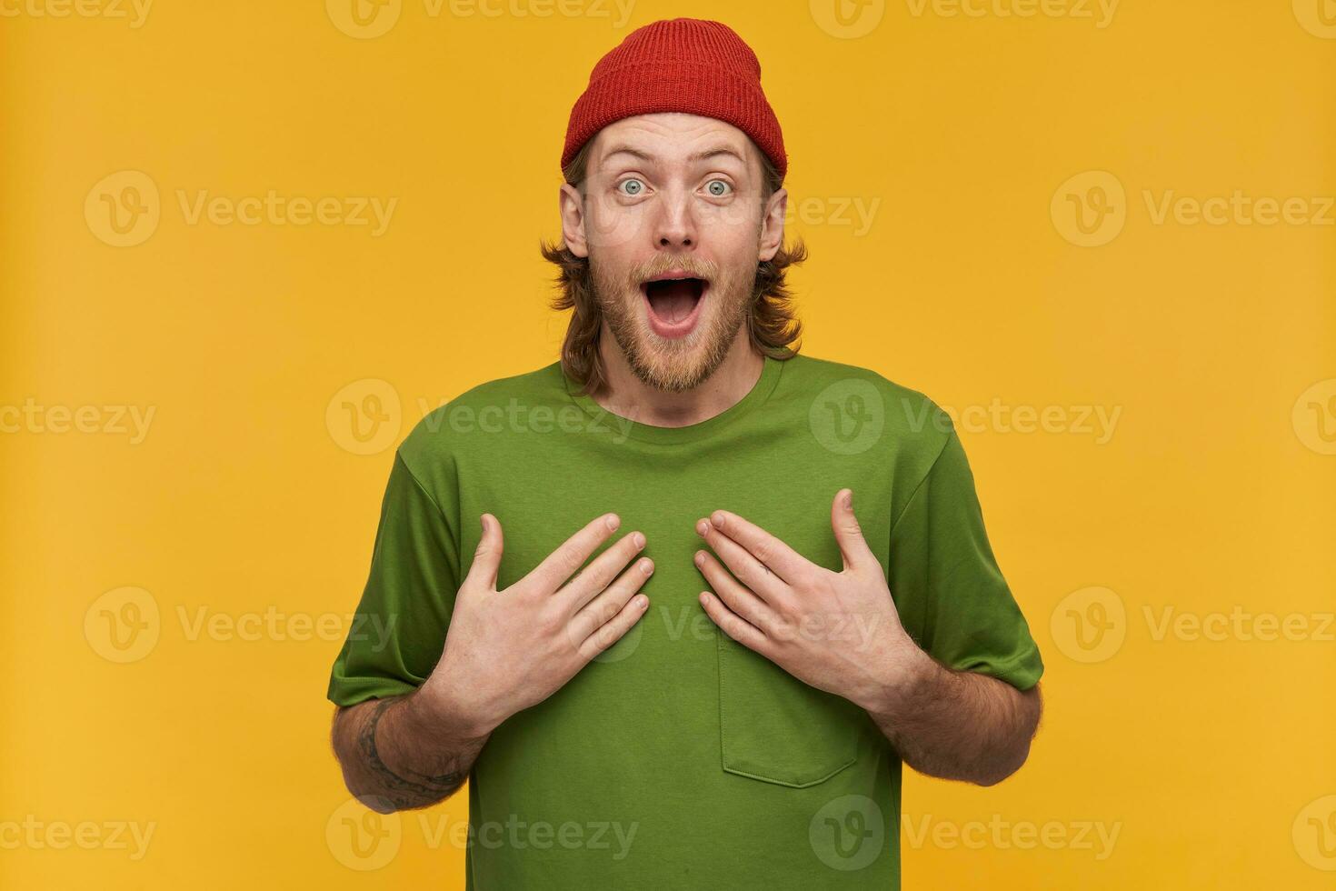 Young man, shocked guy with blond hair, beard and mustache. Wearing green t-shirt and red beanie. Has tattoo. Pointing at himself. Can't believe. Watching at the camera isolated over yellow background photo