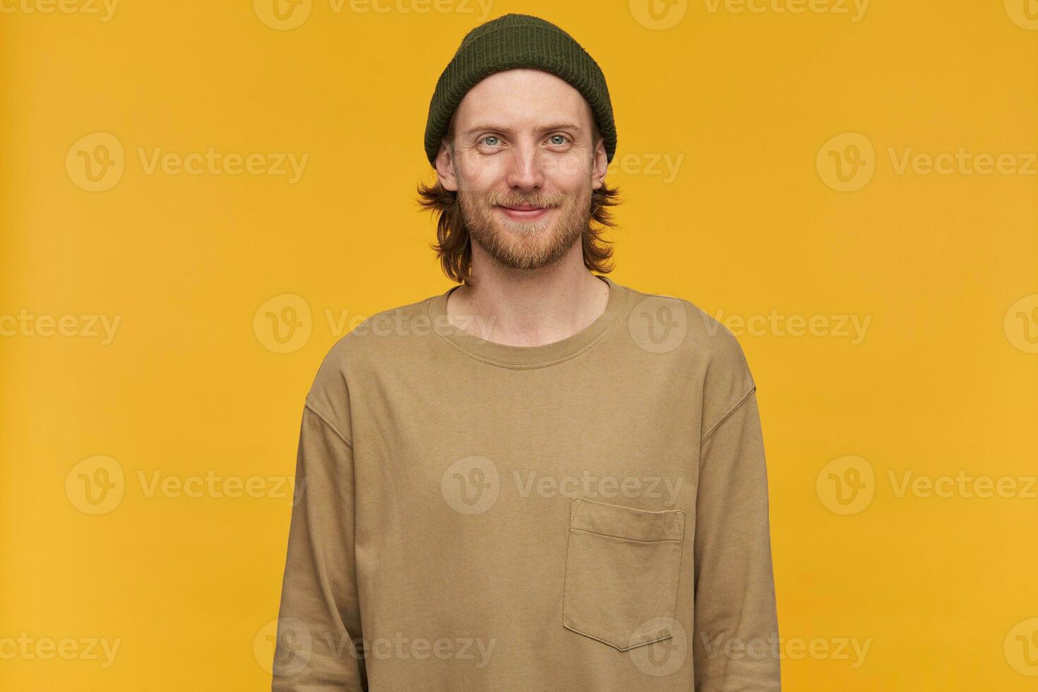 Cool looking male, handsome bearded guy with blond hair. Wearing green beanie and beige sweater. Watching at the camera with confident smile, isolated over yellow background photo
