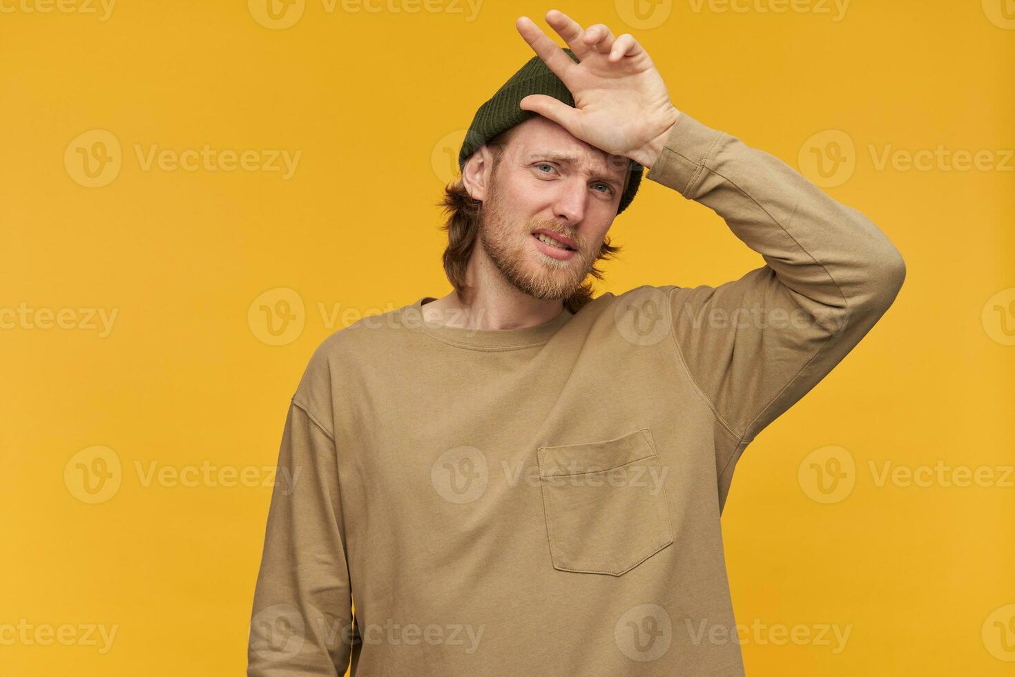 Sick looking male, tired bearded guy with blond hair. Wearing green beanie and beige sweater. Touching forehead with hand. Watching at the camera isolated over yellow background photo