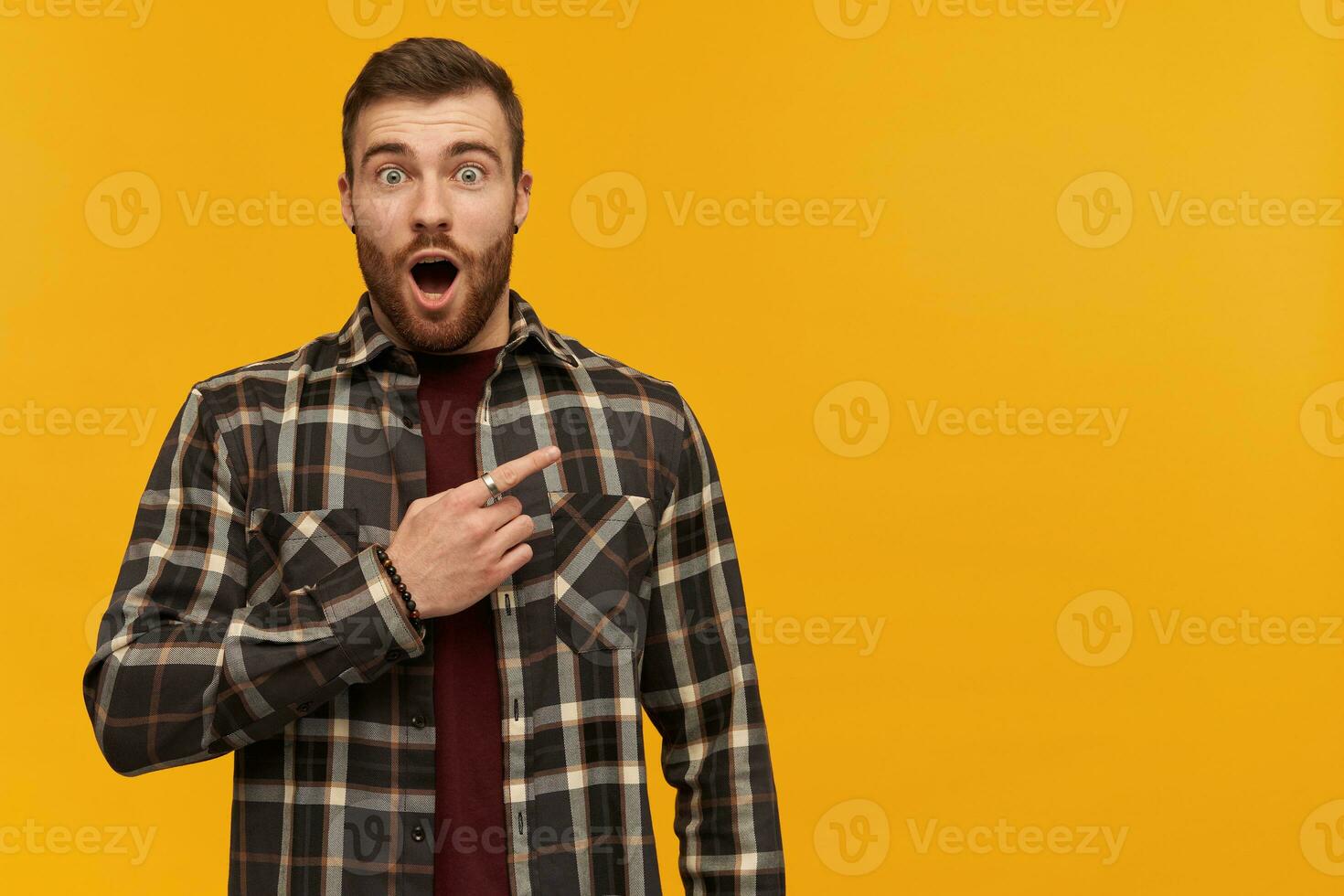 Amazed attractive young bearded man in plaid shirt with opened mouth looks astonished and pointing away to the side over yellow background photo