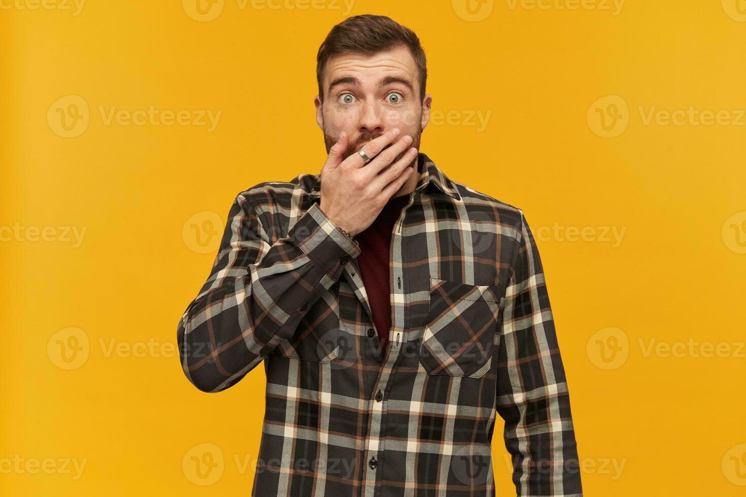 Scared shocked young man in plaid shirt with beard covered his mouth by hand and looks stunned over yellow background photo