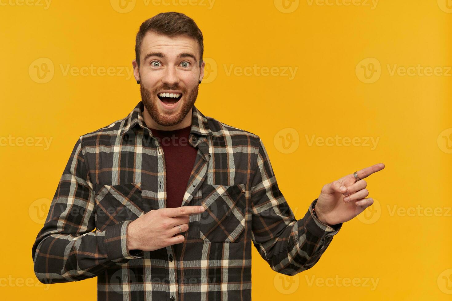 Happy wondered young man in plaid shirt with beard and opened mouth looks amazed and pointing away at empty space by two fingers over yellow background photo