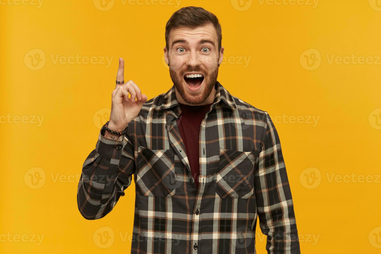 Happy exited young bearded man in plaid shirt shouting pointing up by finger and having an idea isolated over yellow background photo