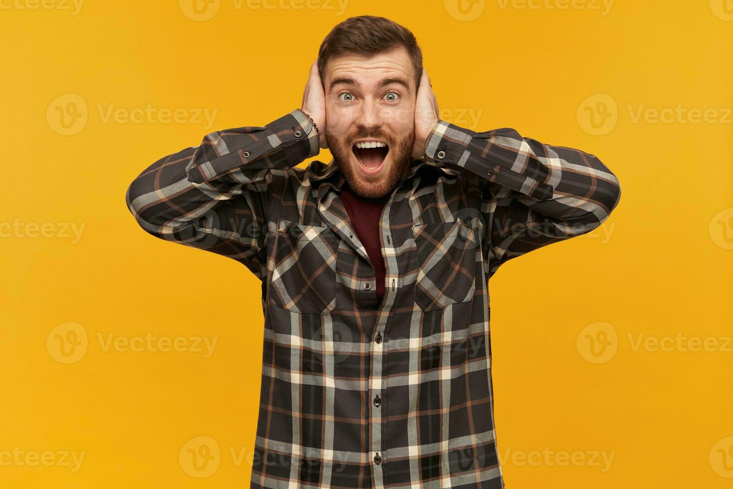 Cheerful excited young man with beard and opened mouth in plaid shirt covered his ears by hands and shouting over yellow background photo