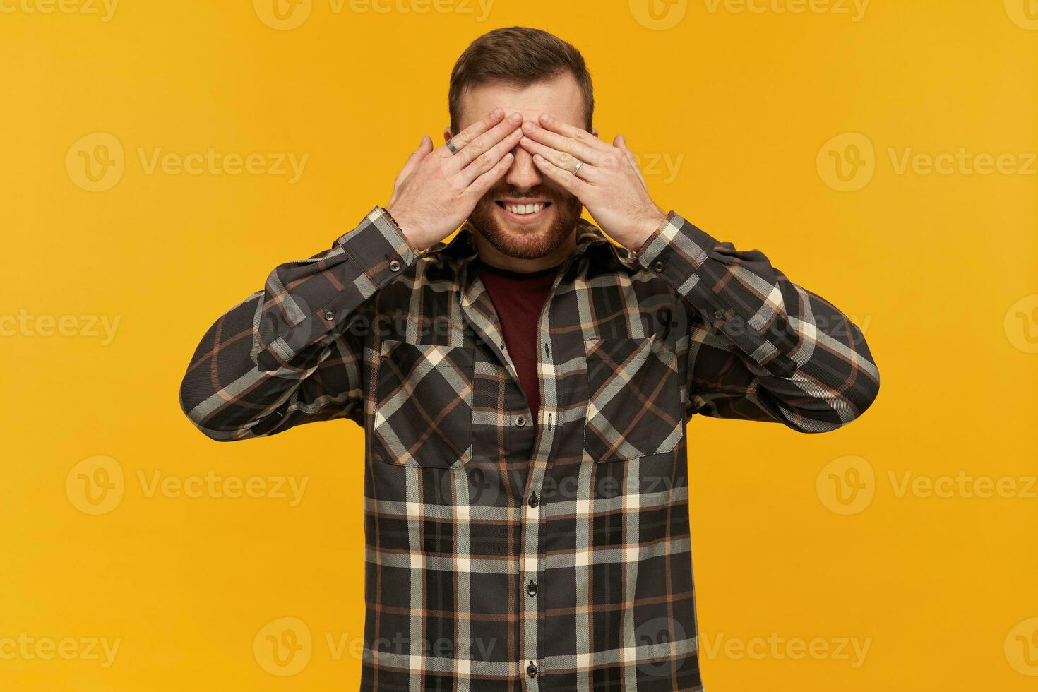 Smiling handsome bearded young man in plaid shirt covered his eyes by hands over yellow background photo
