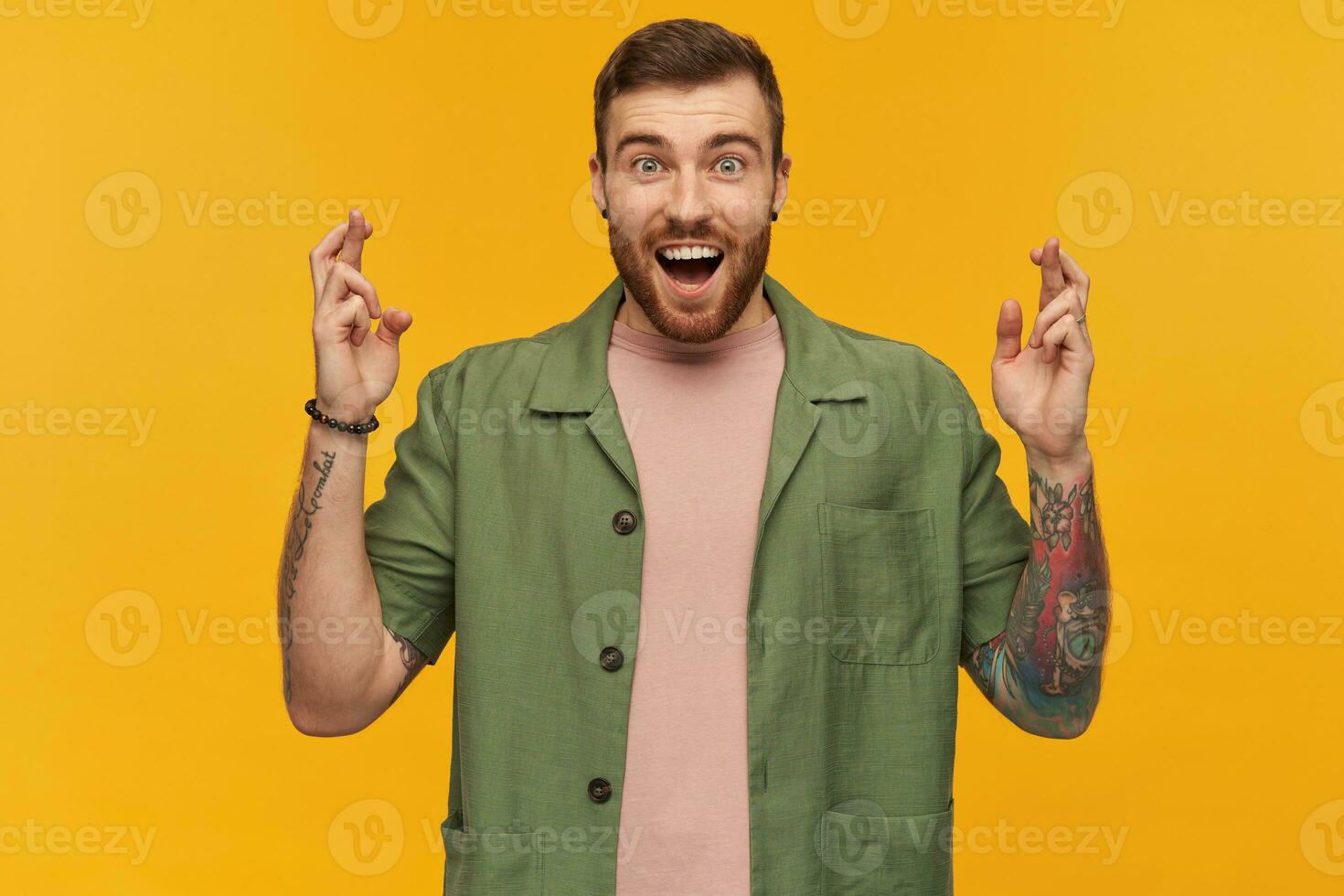 Happy excited young man with beard and tattoo looks inspired keeps fingers crossed and making a wish over yellow background photo