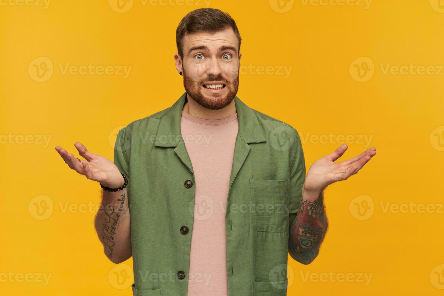 Confused handsome young man with beard and tattoo looks embarrassed and holding copyspace at two both palms over yellow background photo