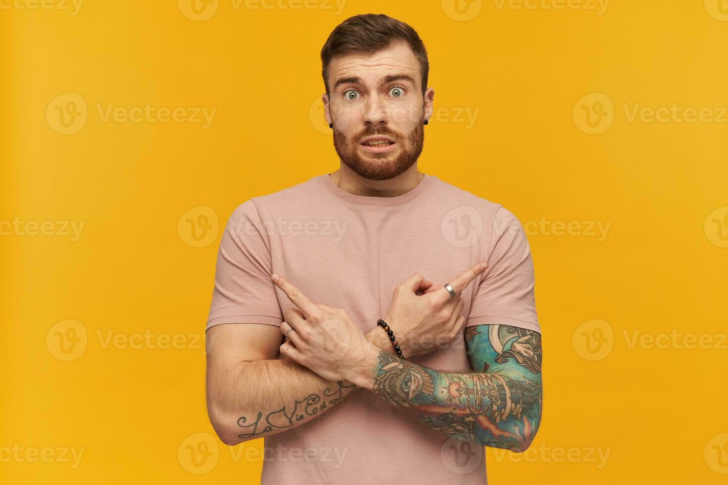 confuso hermoso tatuado joven barbado hombre en rosado t camisa haciendo X forma con brazos y y señalando a ambos lados por dedos terminado amarillo antecedentes foto