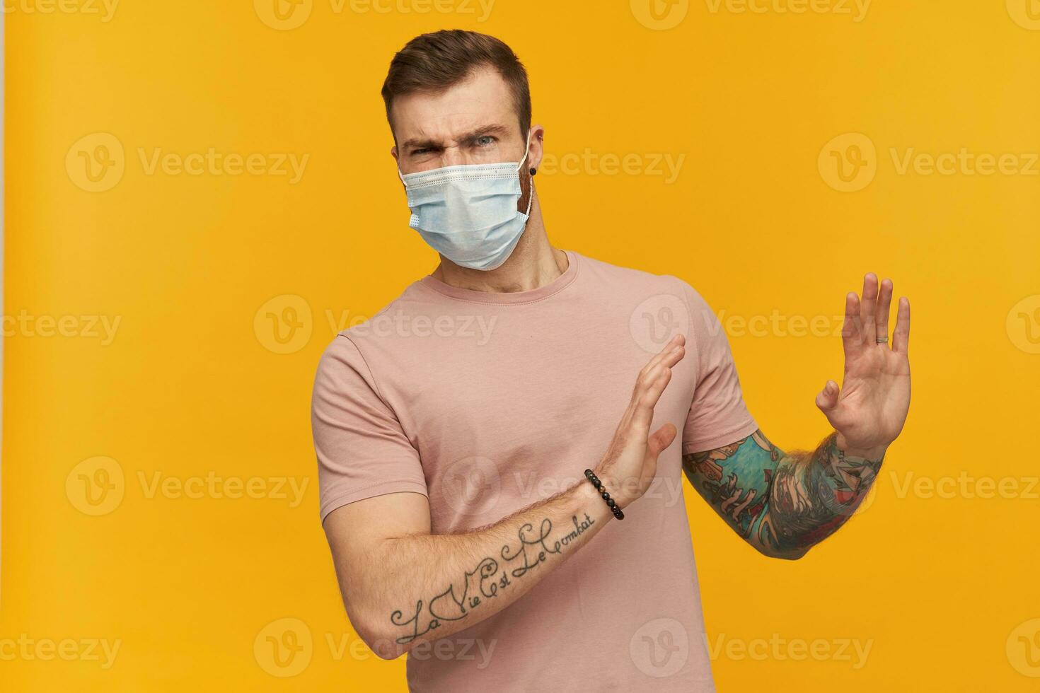 irritado joven barbado hombre con tatuaje en mano en rosado t camisa y higiénico máscara a evitar infección mantiene manos en frente de él mismo defendiendo y rechazando terminado amarillo antecedentes foto