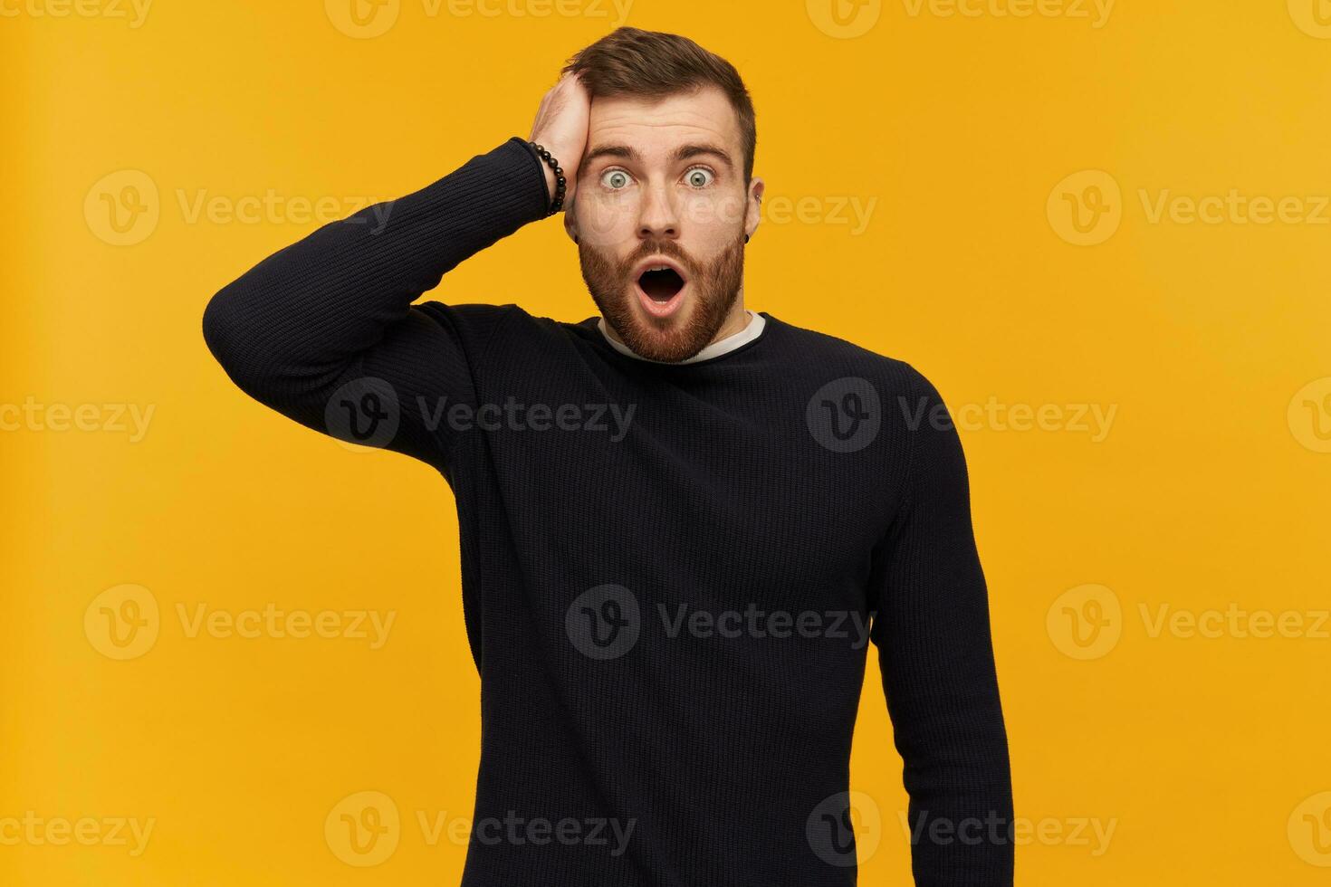 Shocked surprised young man with beard and opened mouth in black long sleeve keeps hand on head and looking at camera over yellow background photo