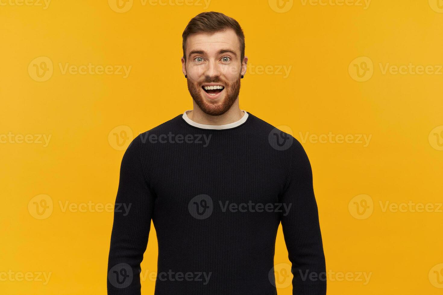 Cheerful amazed young man with beard and opened mouth in black long sleeve feels excited and looks surprised over yellow background photo