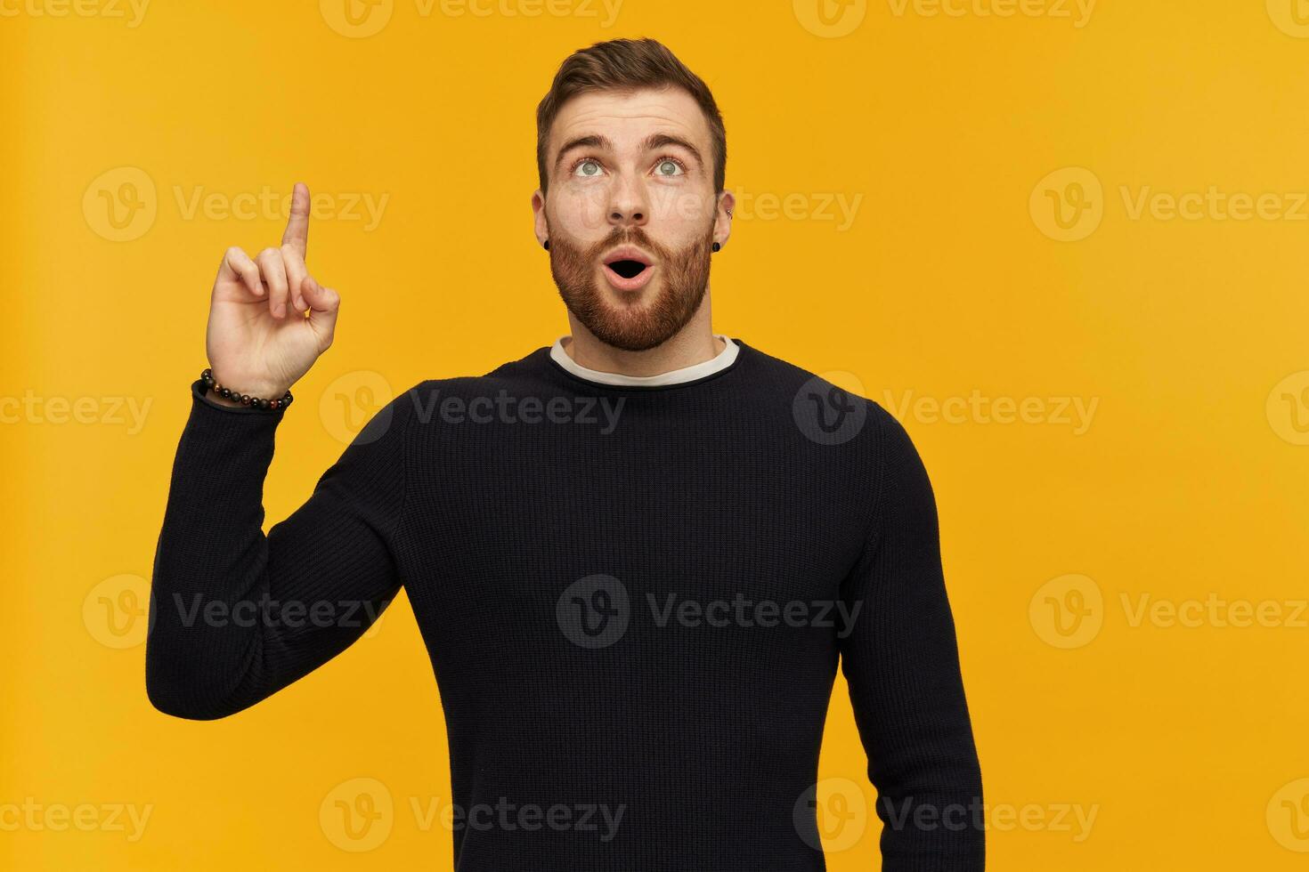 Amazed handsome young man with beard and opened mouth in black long sleeve pointing and looking up to the sky over yellow background photo
