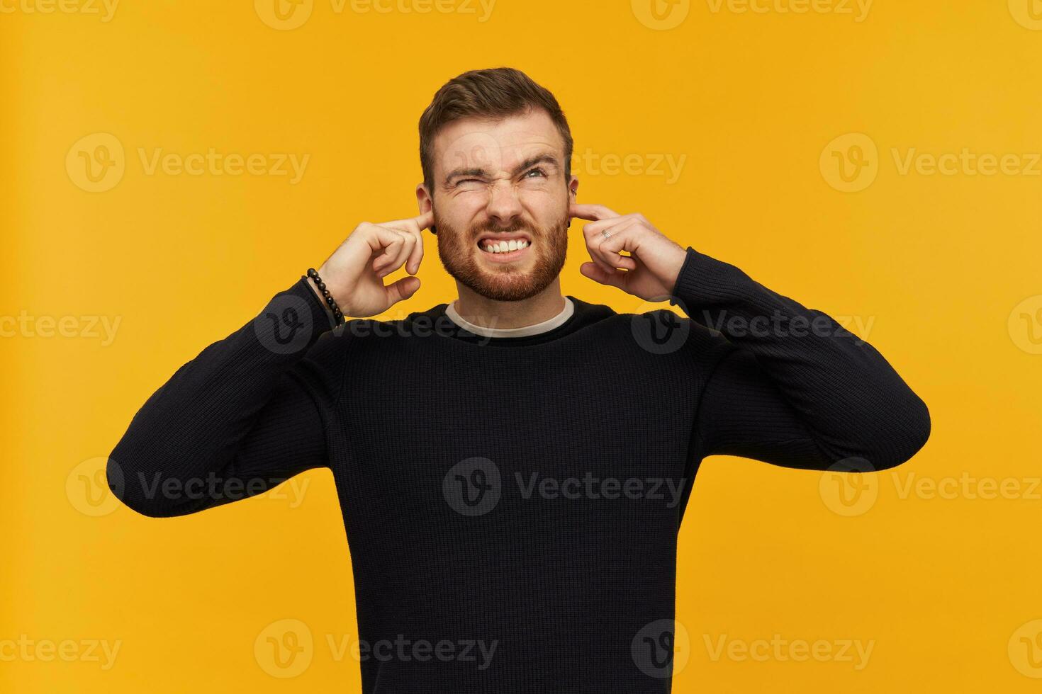 irritado enojado joven hombre con barba en negro largo manga mira irritado y cerrado orejas por dedos en pie terminado amarillo antecedentes foto