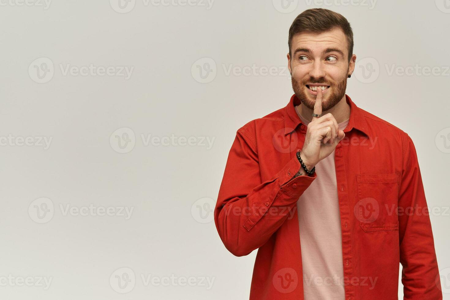 Pensive confused young bearded man in red shirt showing silence gesture and looking to the side away over white background photo