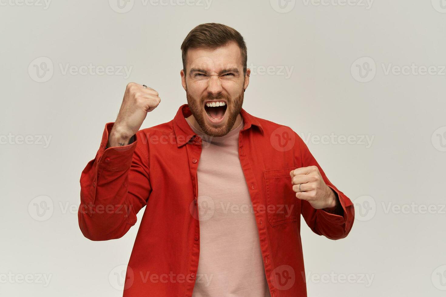 Cheerful excited young man in red shirt with beard keeps fists clenched and raised hands and screaming over white background Celebrating victory concept photo