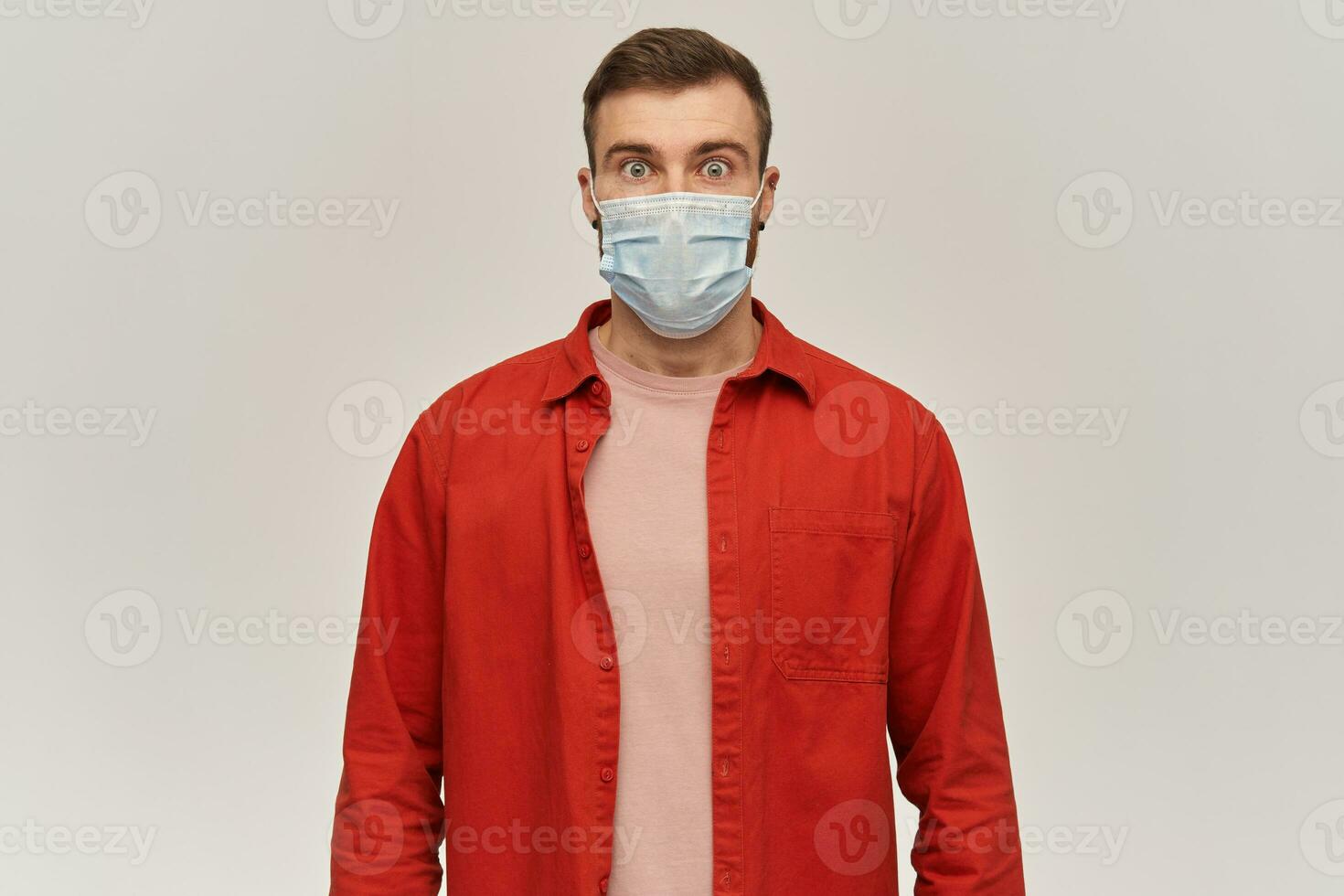 Worried shocked young bearded man in red shirt and virus protective mask on face against corona virus standing and looking at camera over white background photo