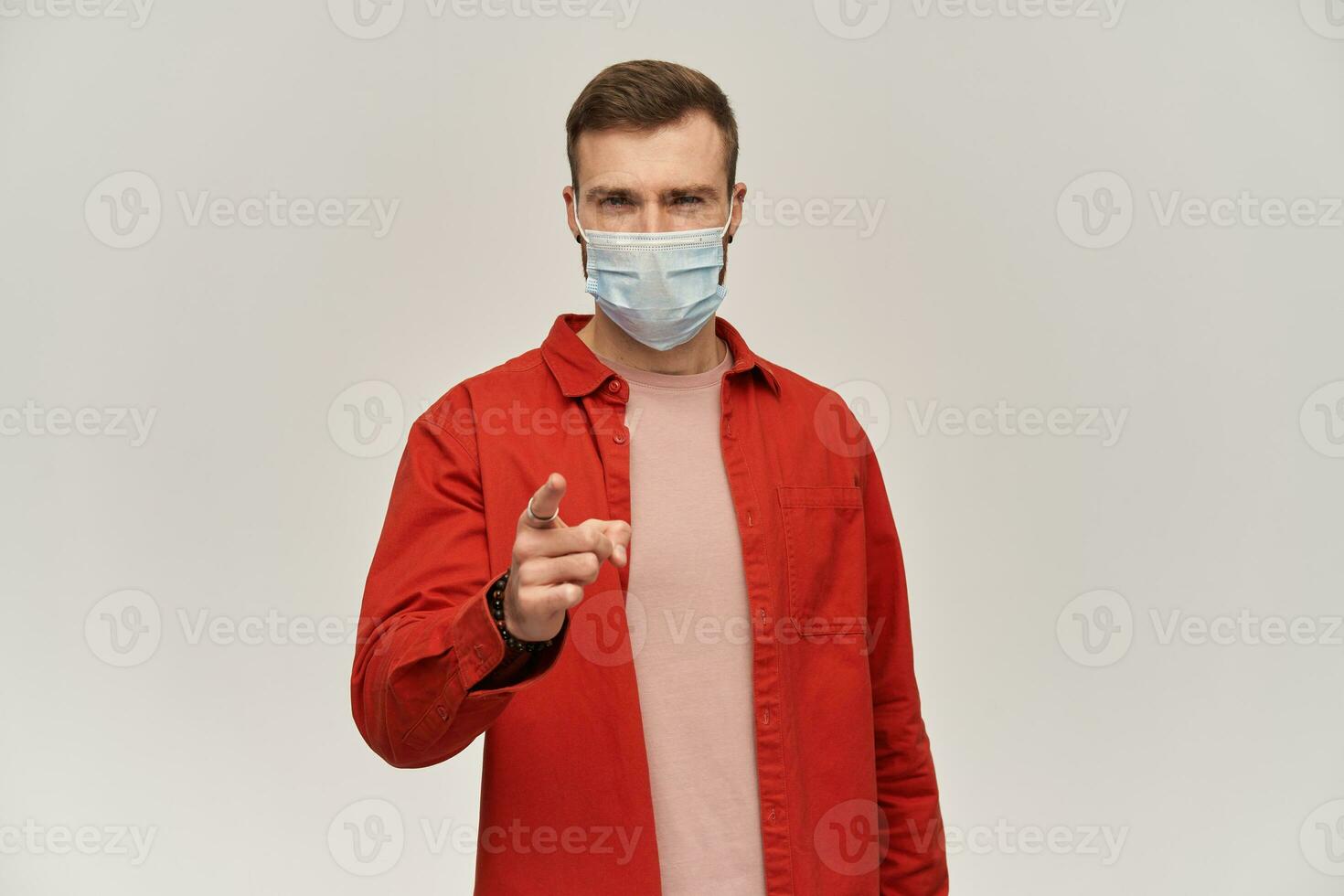 Skeptical young man with beard in red shirt and hygienic mask to prevent infection pointing on camera or you by finger over white background photo