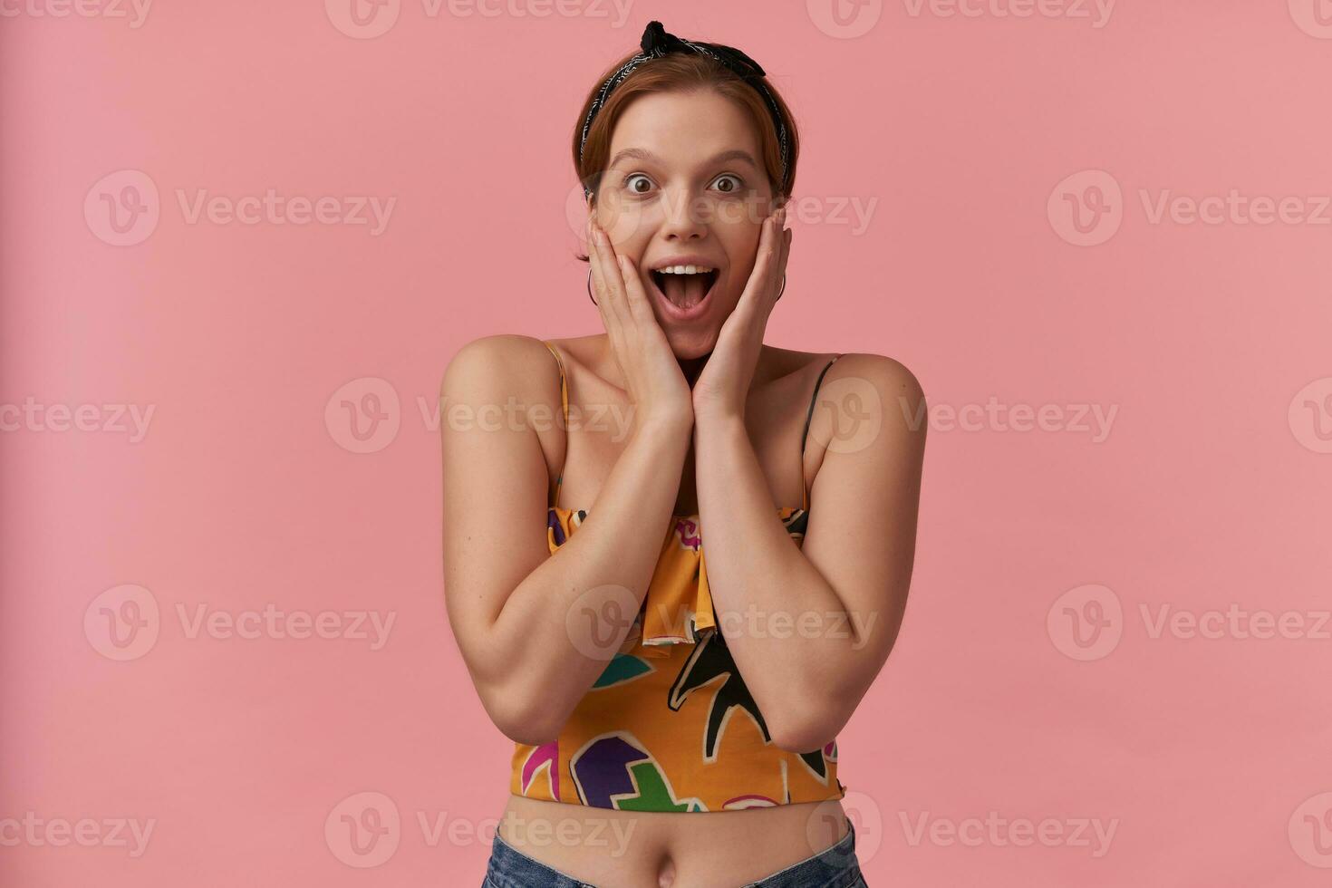 Portrait of european charming woman 20s with natural makeup wearing stylish summer crop top slim strap and bandana surprised emotion with arms touching face isolated posing against pink background photo
