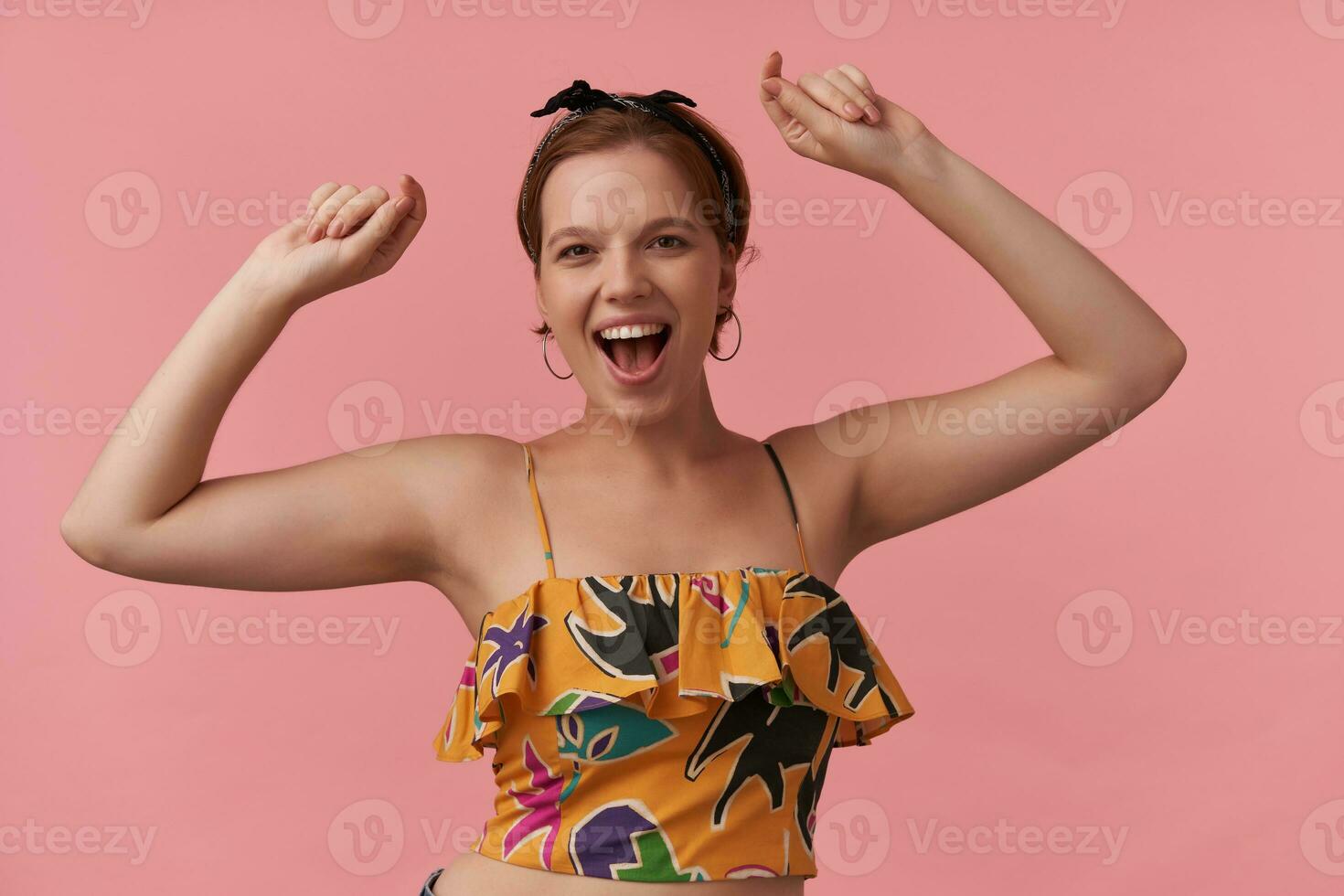 Portrait attractive young brown eyed with natural makeup wearing summer style colorful outfit ear rings and bandana smiling at you against pink wall with arms up emotion glad happy dancing celebrating photo