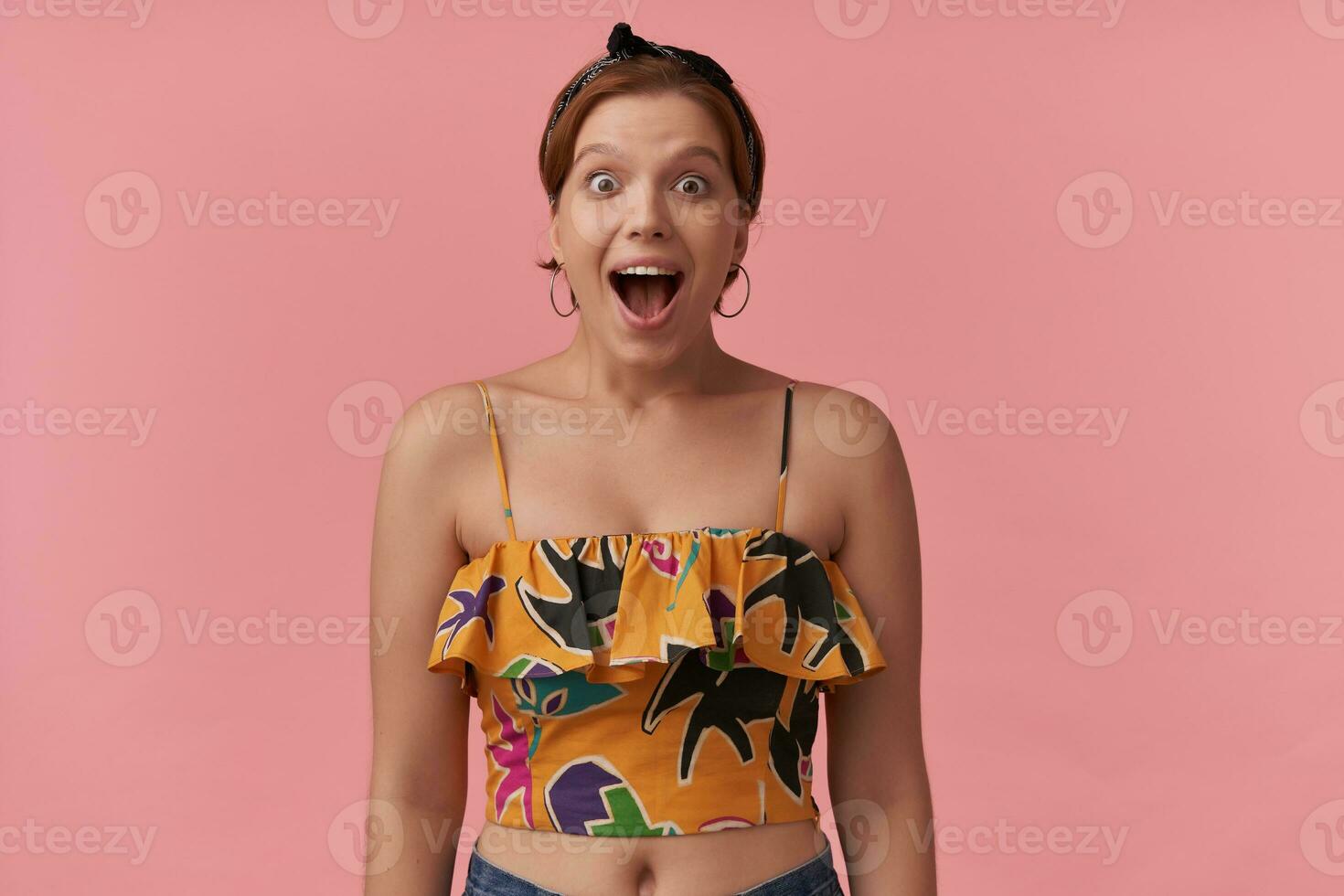 Portrait of 20s european woman wearing summer style yellow crop top and bandana surprised happy excited emotion face natural makeup brown eyes looking at you isolated over pink background photo