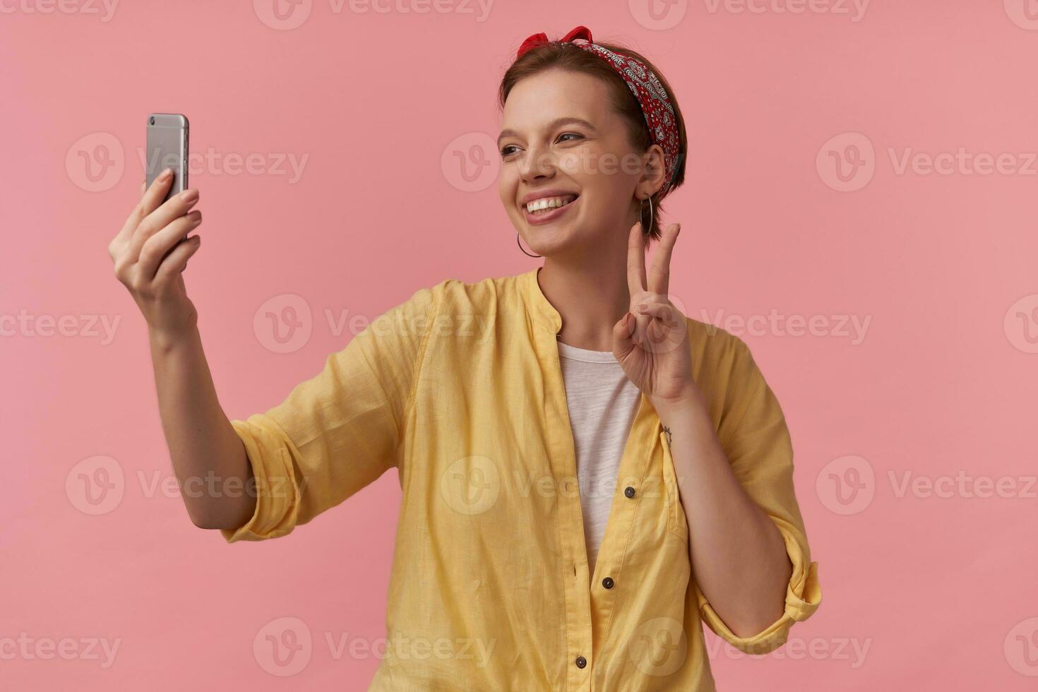 Image of european charming woman 20s with natural makeup wearing trendy summer posing over pink background emotion happy glad smiling making self shot two fingers up arms gesticulates on the phone photo