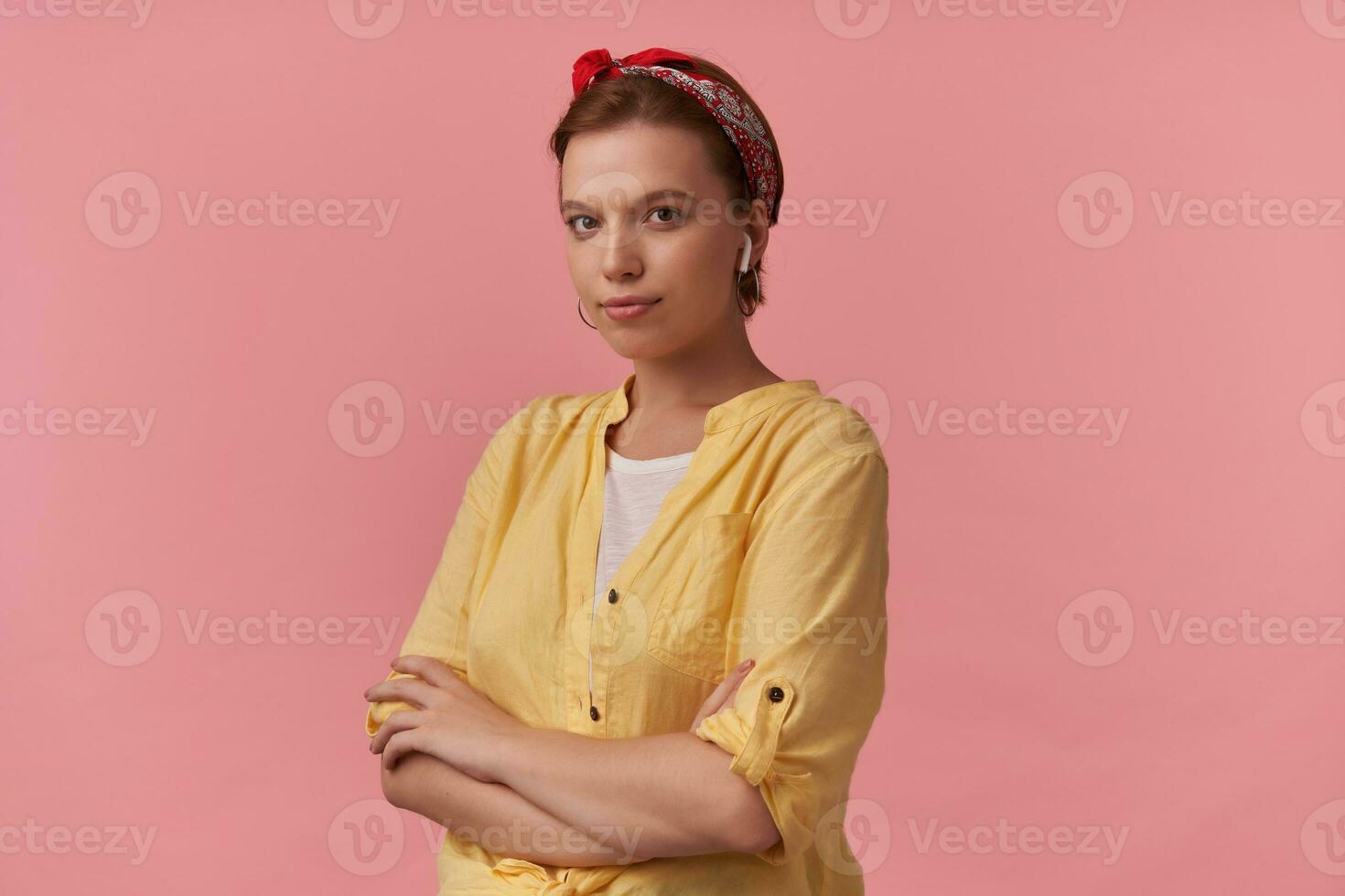 Portrait of attractive young brown eyed charming woman 20s with natural makeup wearing white t-shirt and yellow shirt and red bandana posing against pink wall emotion looking at you with arms crossed photo