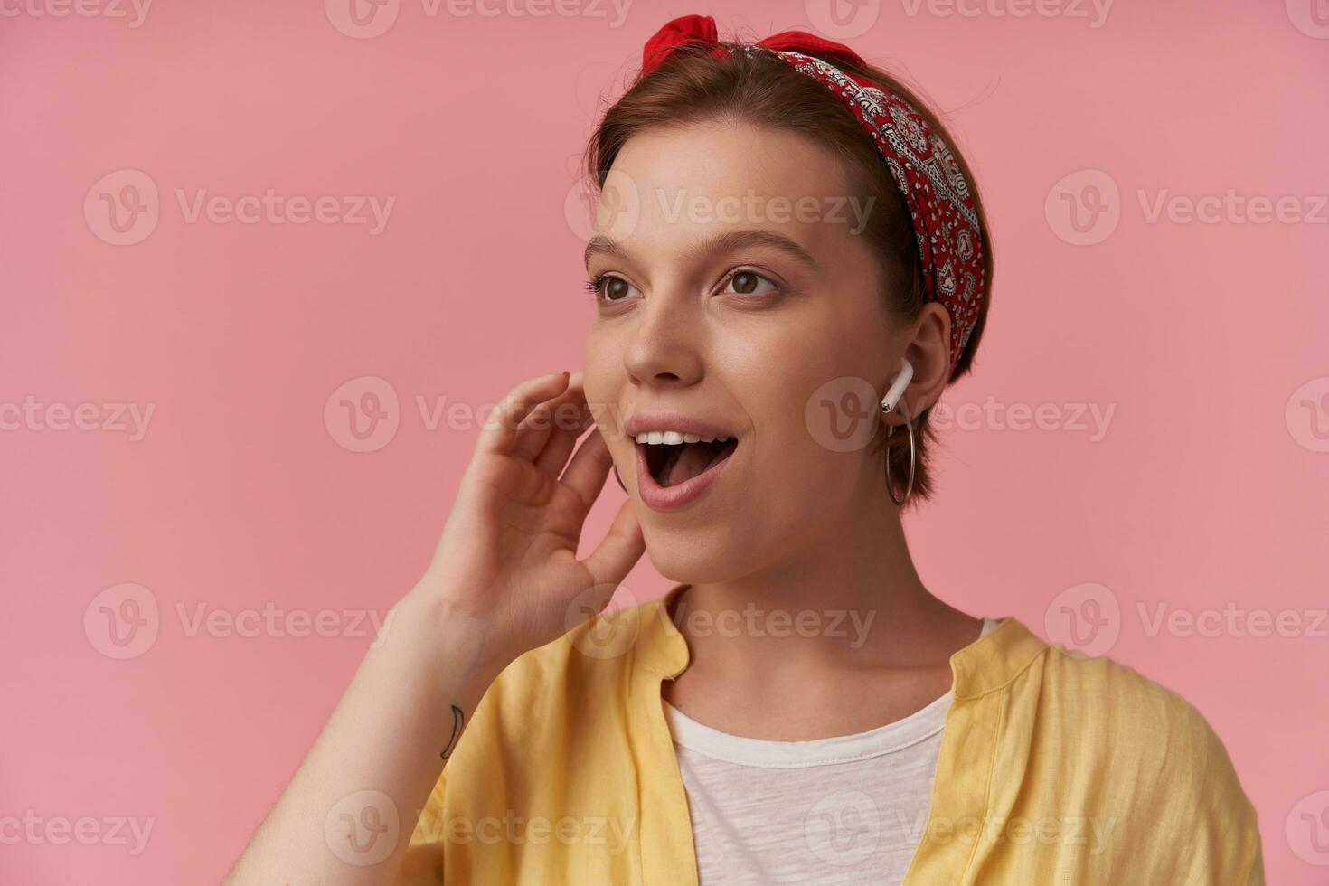 Portrait of young brown eyed european brown haired charming woman 20s with natural makeup wearing white t-shirt and yellow shirt and red bandana emotion looking aside amazed smiling against pink wall photo