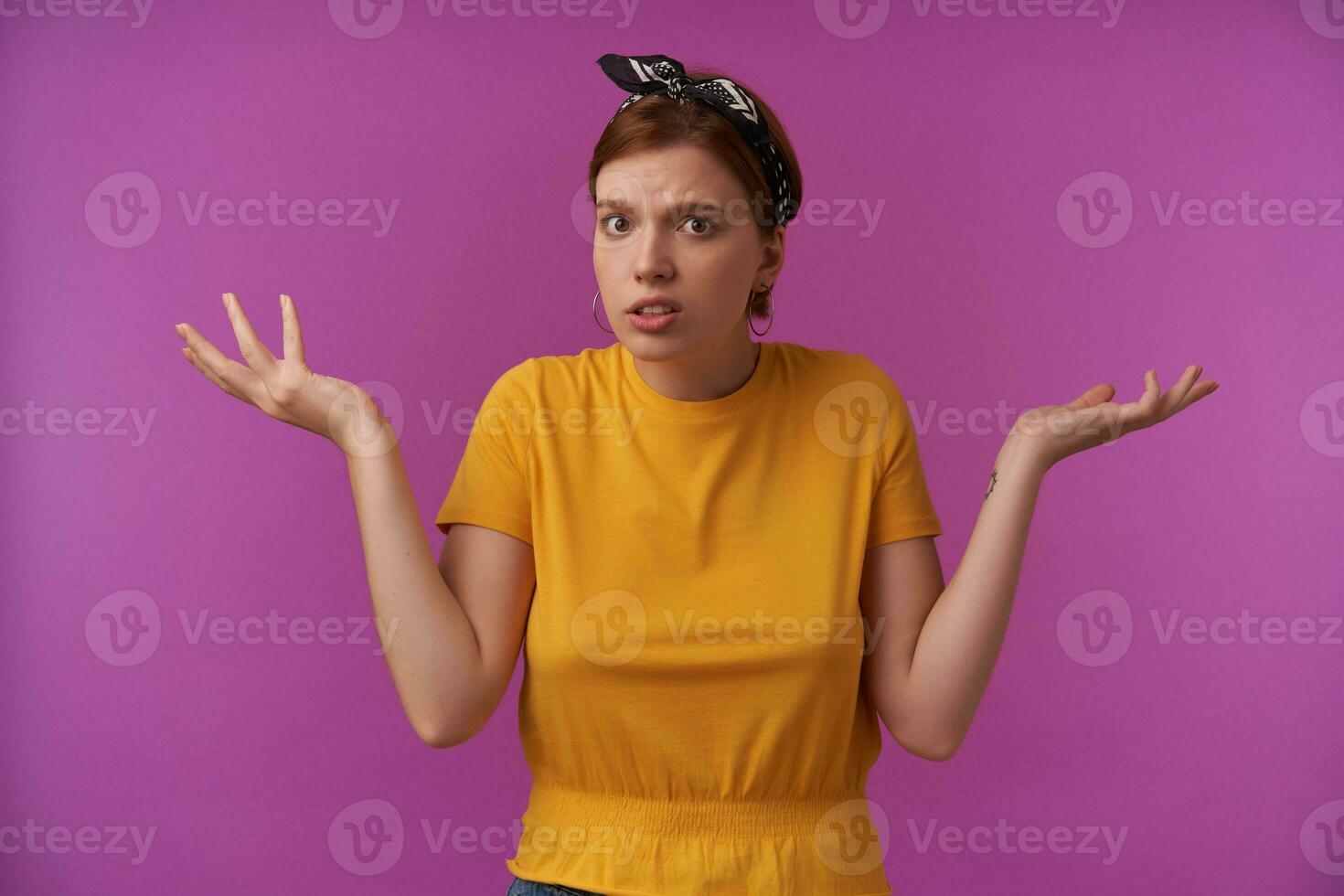 Portrait of attractive young brown eyed european woman with natural makeup emotion confused guess with arms aside wearing stylish yellow t-shirt and black bandana posing against purple background photo