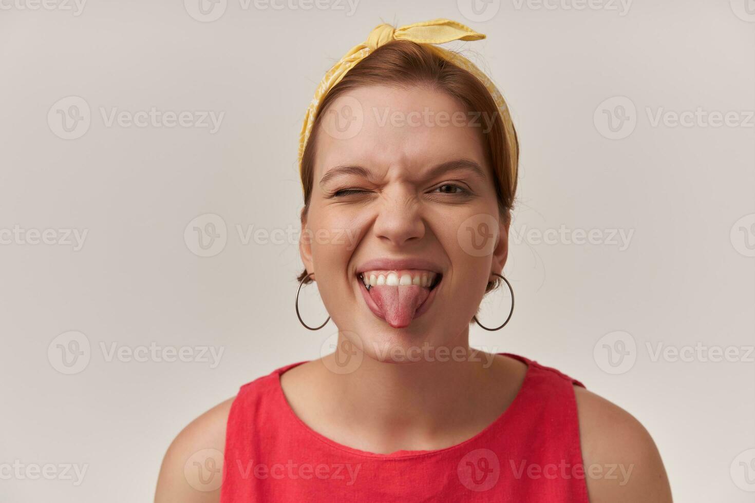 Active young brown-eyed European woman with natural makeup and earrings emotion glad silly smiling at you and wink shoe tongue wearing stylish trendy red dress and yellow bandana over white wall photo