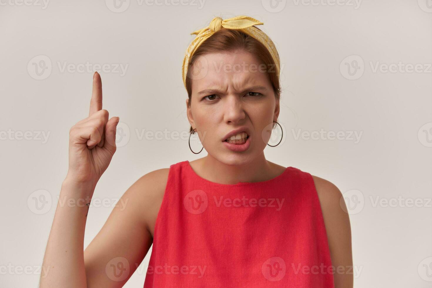 Portrait attractive young brown eyed woman 20s wearing stylish trendy red dress ear rings and yellow bandana posing against white wall with finger point up emotion attention look up haughty arrogant photo