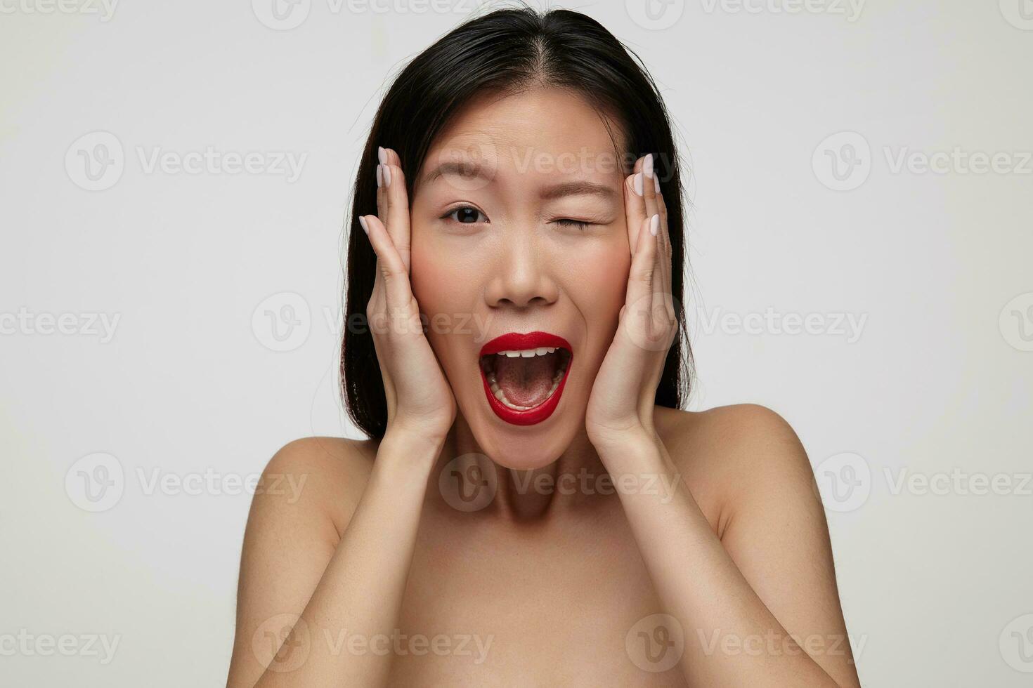 Indoor photo of young agitated dark haired woman holding her face with raised palms while looking excitedly at camera with closed eye, standing over white background