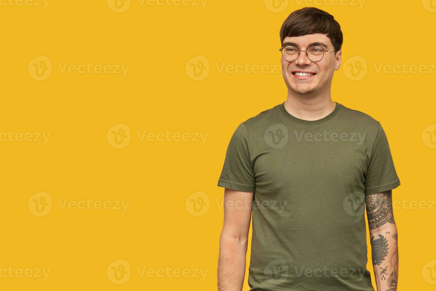 Indoor shot of young attractive male, in round stylish glasses and wears green t-shirt, smiles broadly with happy facial expression. isolated over yellow background. photo