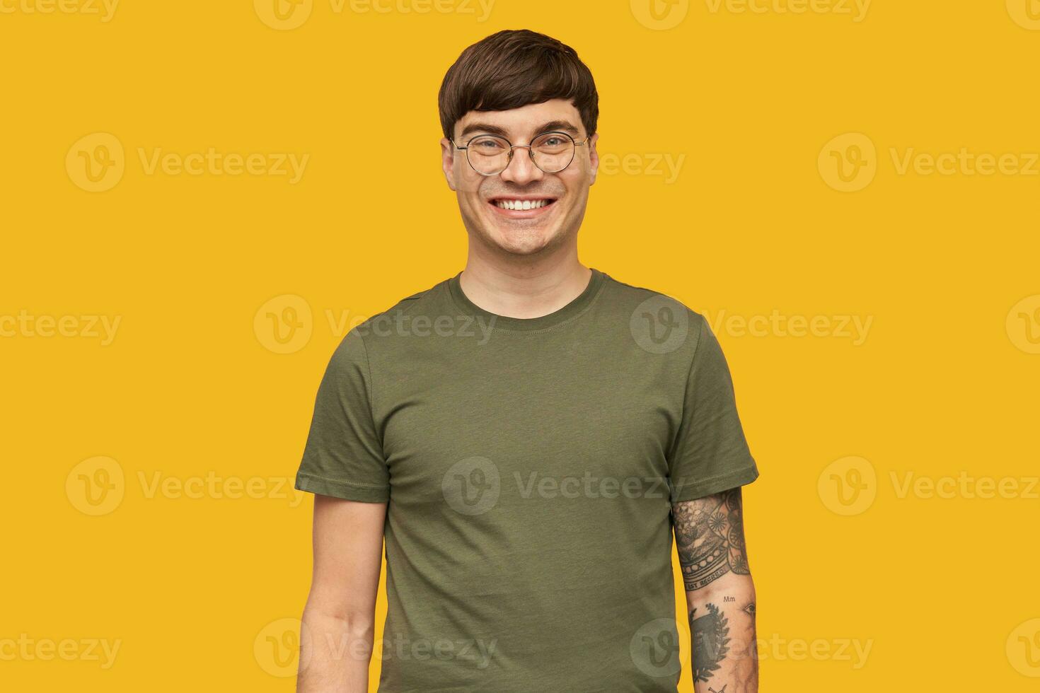 Indoor shot of young attractive male, in round stylish glasses and wears green t-shirt, smiles broadly with happy facial expression. isolated over yellow background. photo